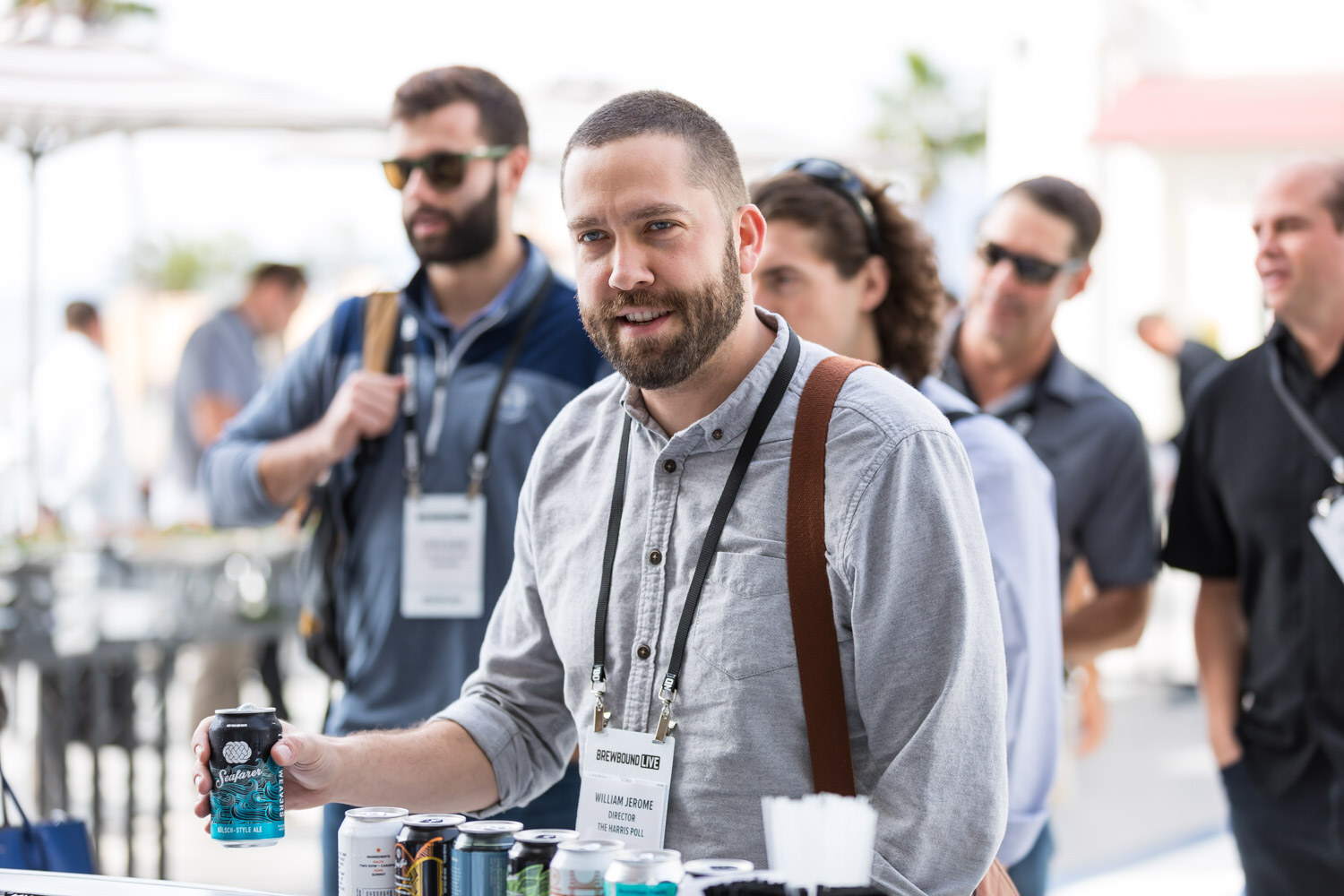A bearded man with a lanyard holds a can of beer, with other attendees in sunglasses behind him.