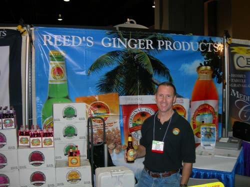 A smiling man in a black polo with a badge holds a bottle in front of a Reed's Ginger Products banner, with cases of ginger beer stacked beside him.