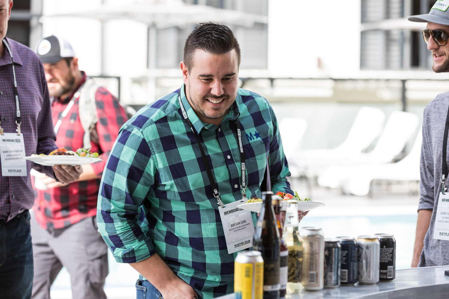 A man in a teal buffalo-plaid shirt holds a plate of food beside a bar of canned beers.