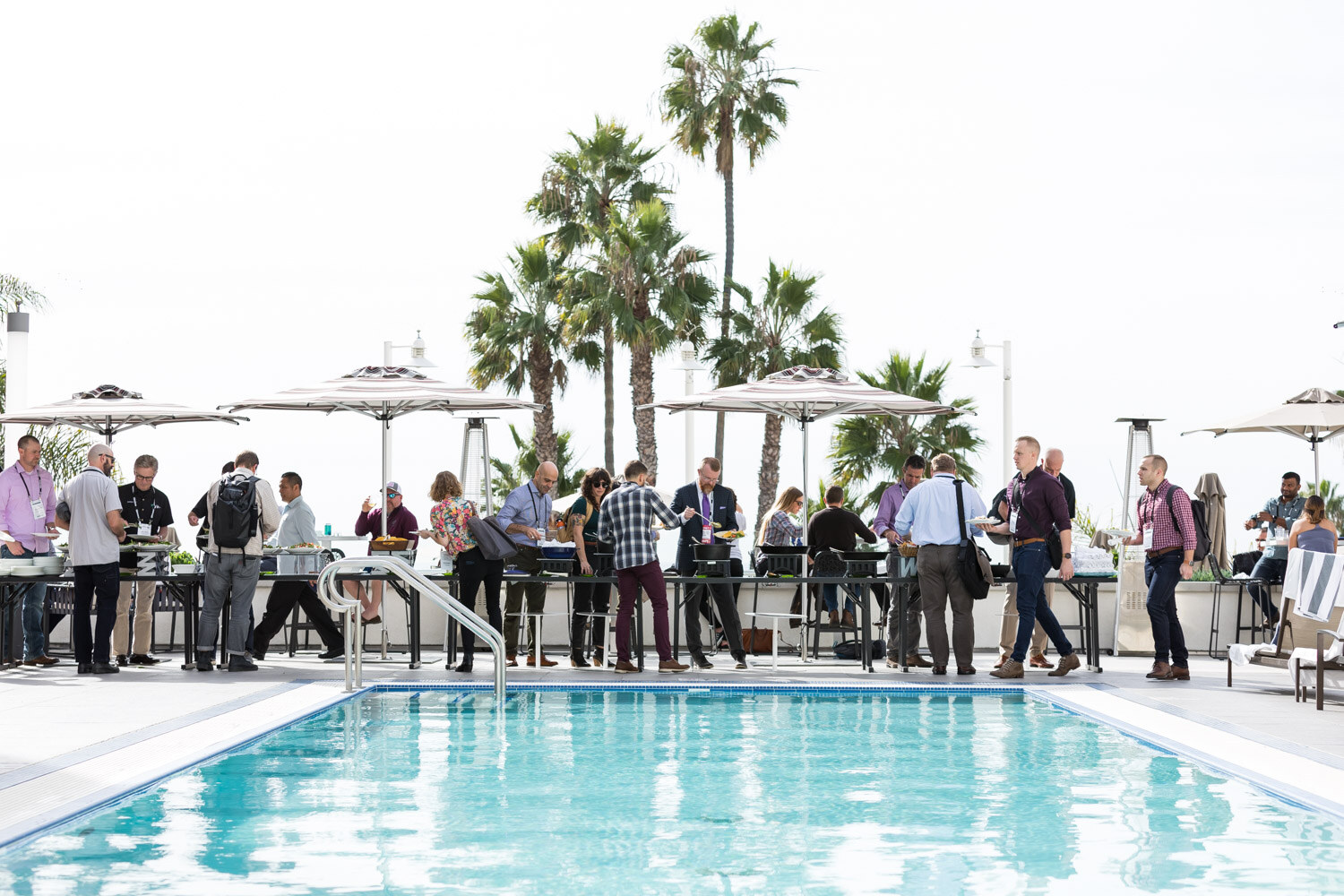 A row of people at a poolside buffet under umbrellas with palm trees and a blue pool in front.