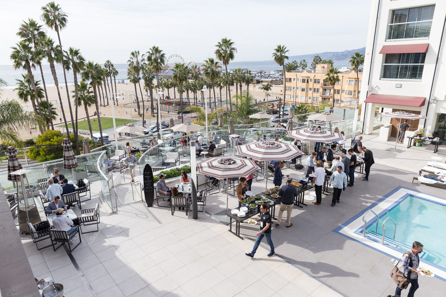 An overhead view of a hotel terrace with striped umbrellas, a pool, palm trees and the ocean in the distance.