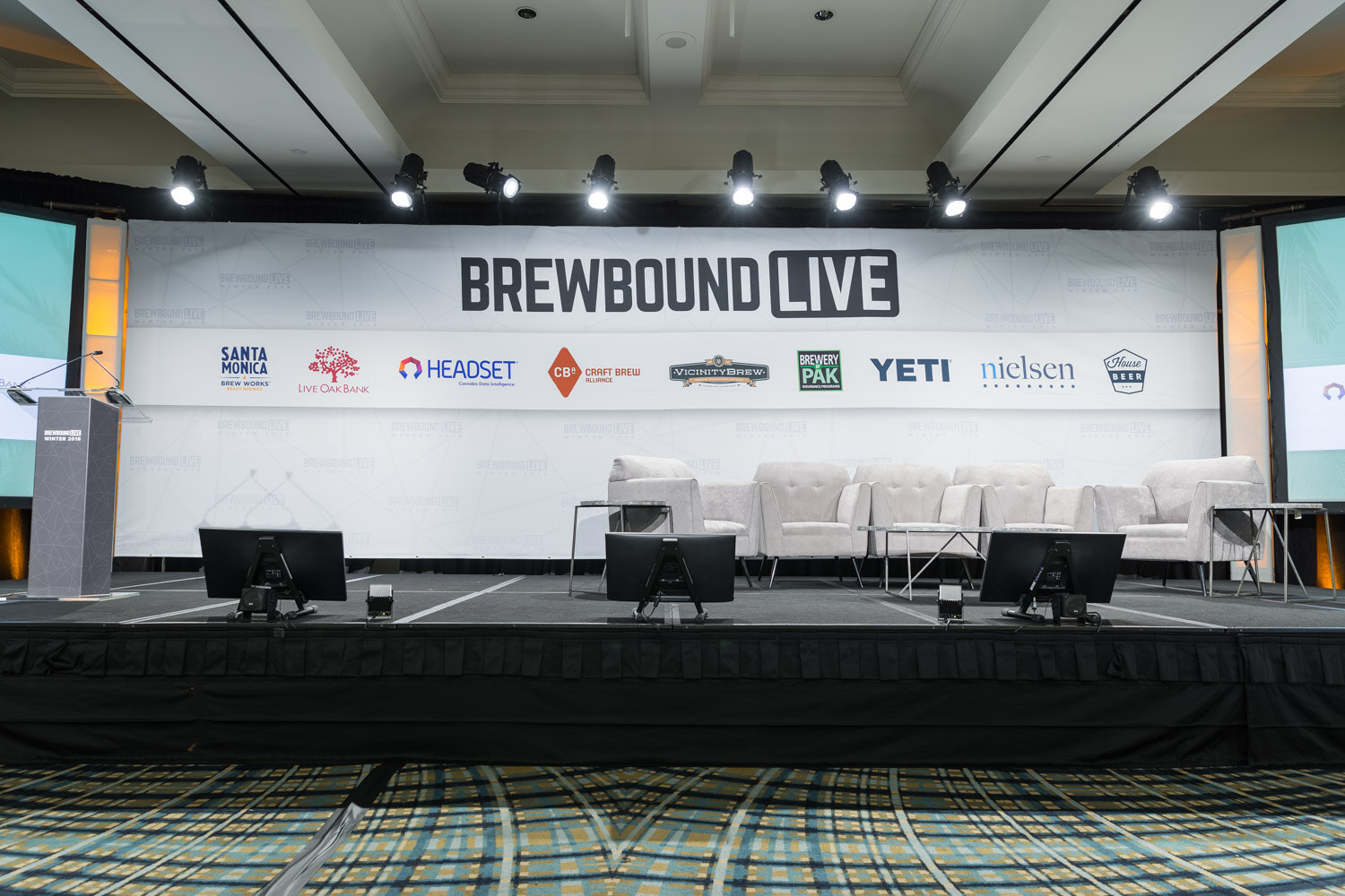 An empty stage with white couches and a podium in front of a Brewbound Live backdrop with sponsor logos including Headset, YETI and Nielsen.