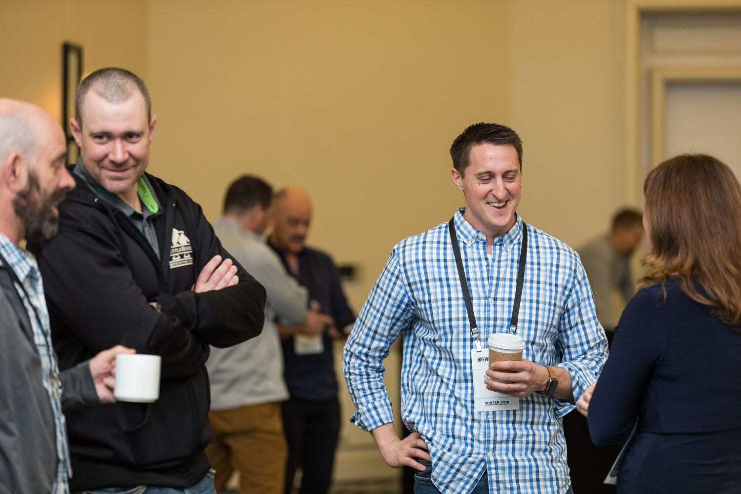 Several attendees with lanyards and coffee cups talk and laugh in a hotel hallway.