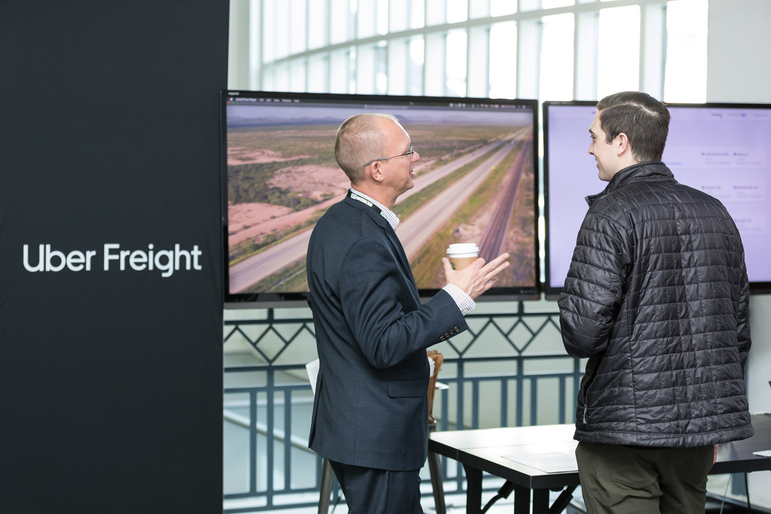 Two men stand in front of a screen with an aerial map next to a black sign reading Uber Freight.