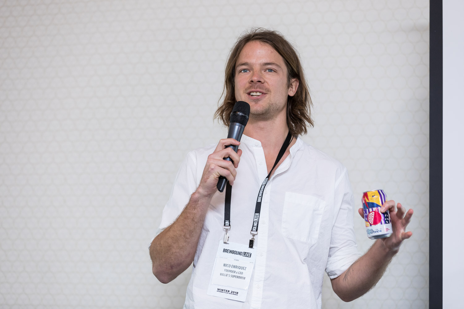 A man with long hair in a white shirt holds a microphone in one hand and a colorful can in the other.