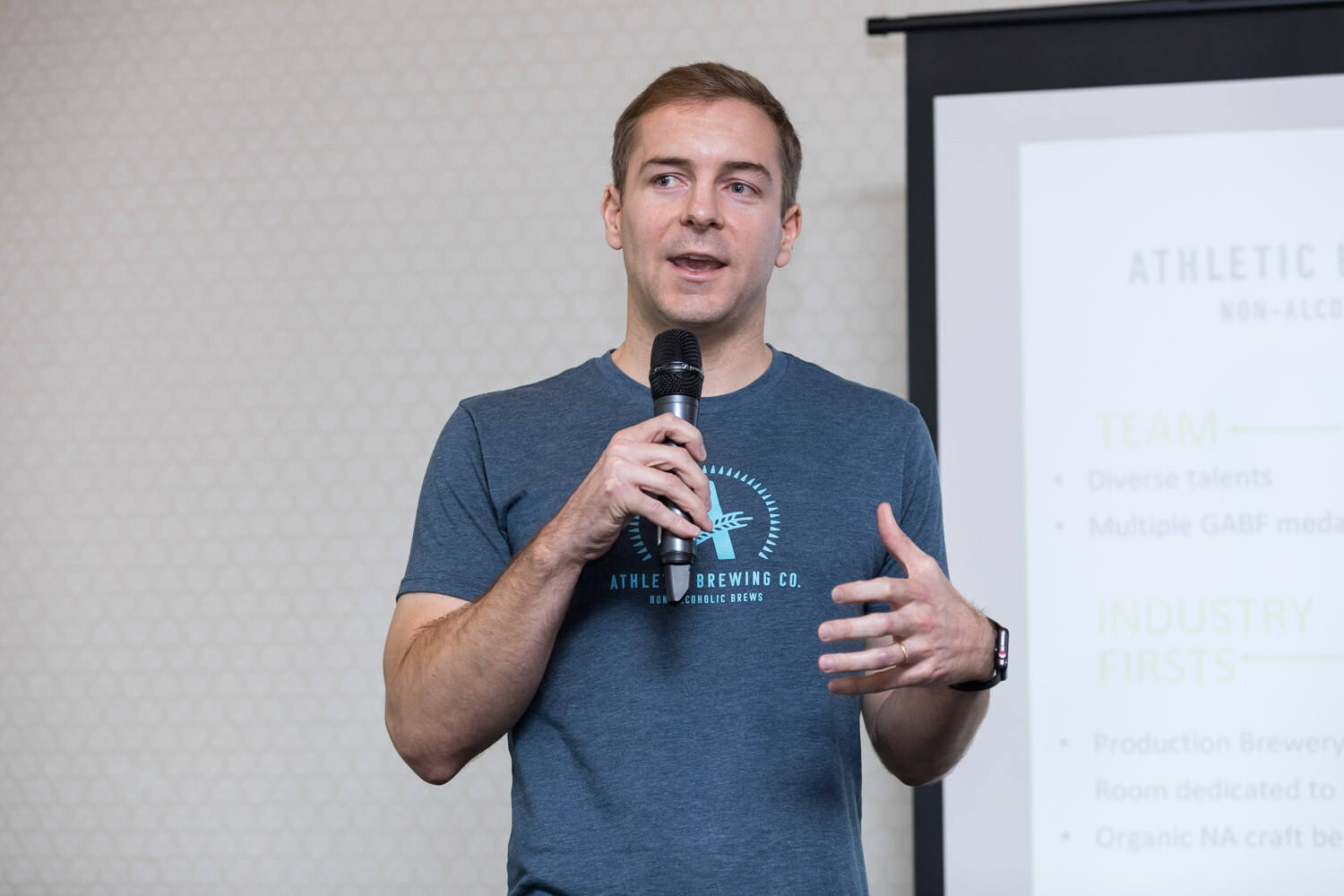 A man in a blue T-shirt reading Athletic Brewing Co. speaks into a microphone beside a projection screen.