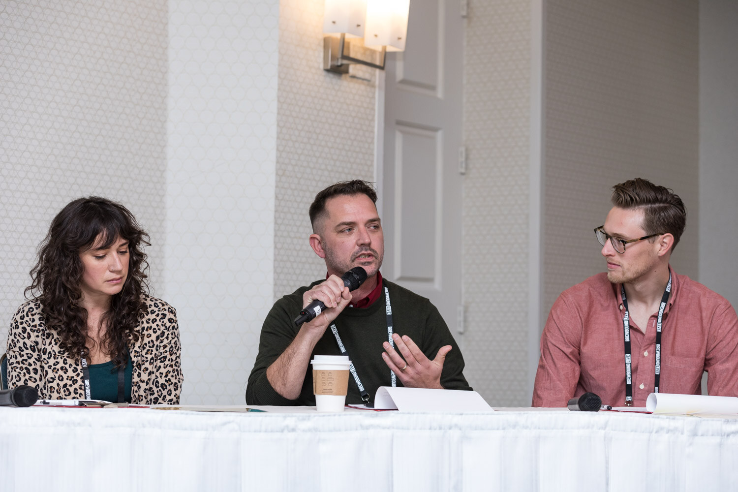 Three people seated behind a table with a white cloth, the middle one speaking into a microphone.