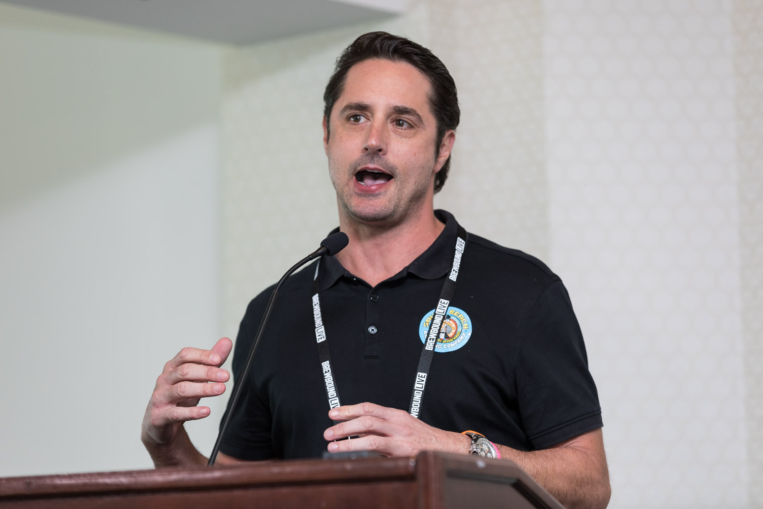 A man in a black polo shirt and lanyard speaks at a wooden podium in a hotel meeting room.