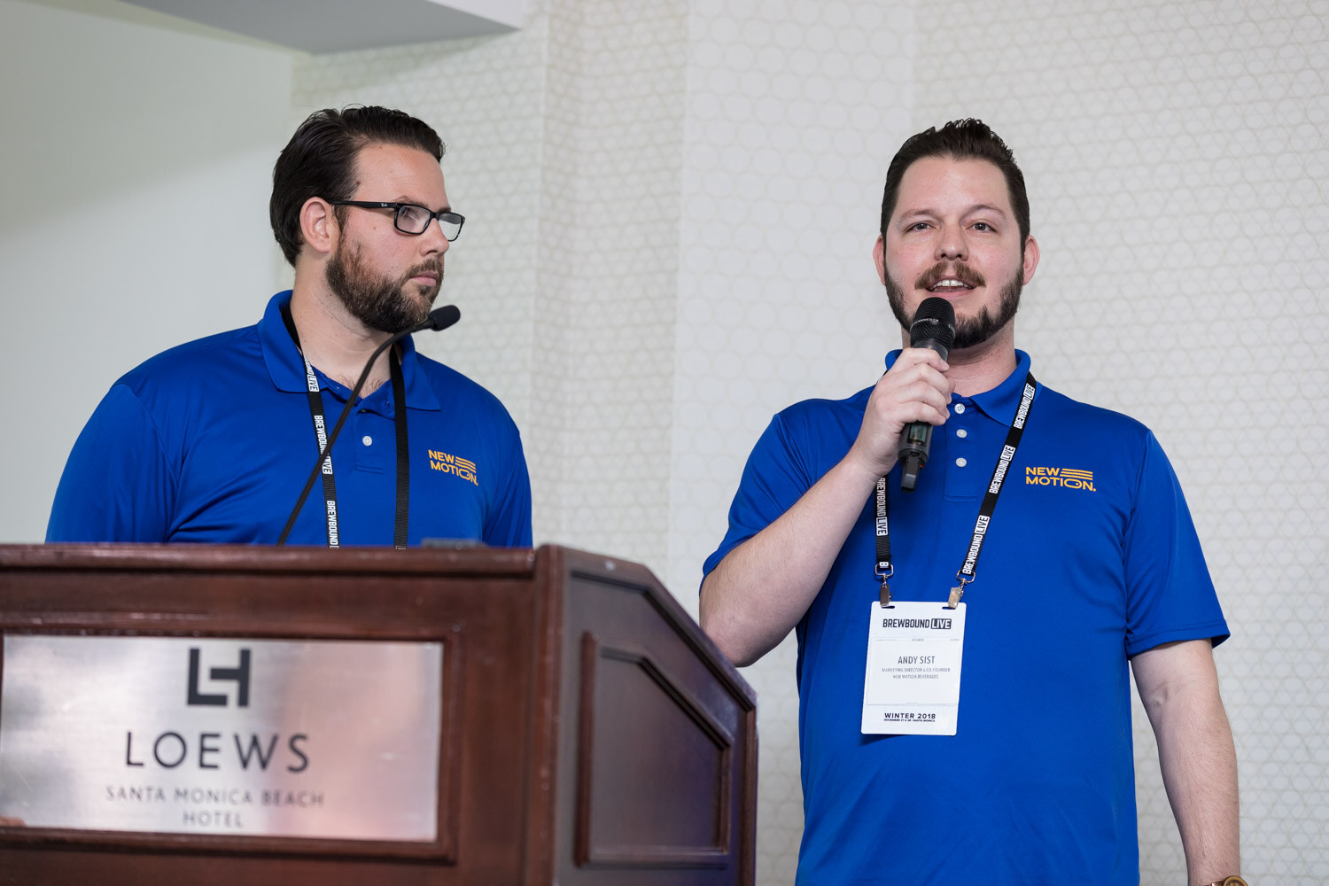 Two men in royal blue polo shirts and lanyards stand at a podium marked Loews Santa Monica Beach Hotel, one holding a microphone.