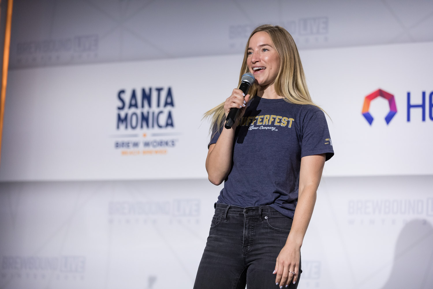 A woman in a navy Sufferfest T-shirt holds a microphone on stage in front of a Santa Monica Brew Works logo.