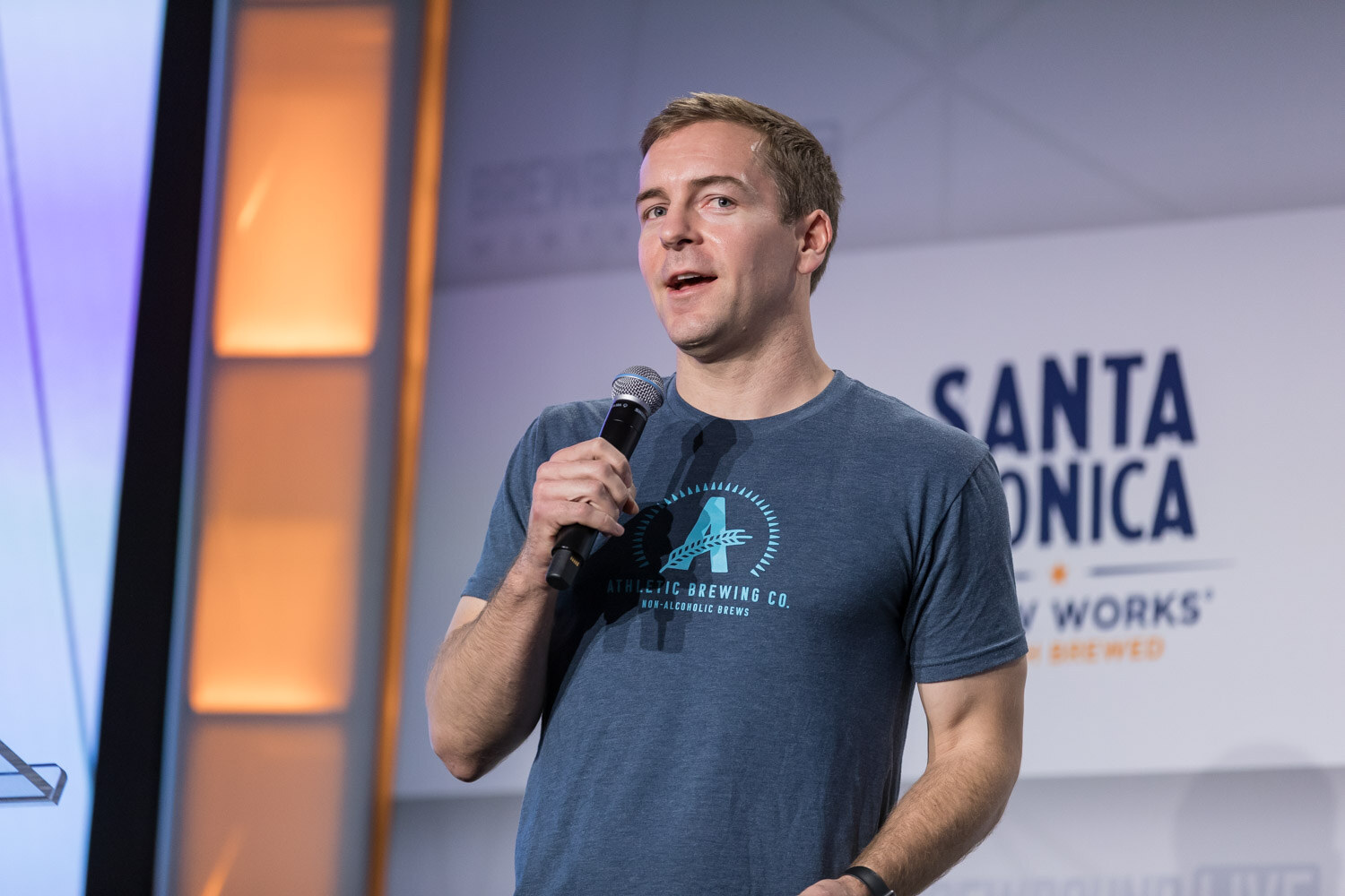 A man in a blue Athletic Brewing Co. T-shirt holds a microphone on stage in front of a Santa Monica Brew Works logo.