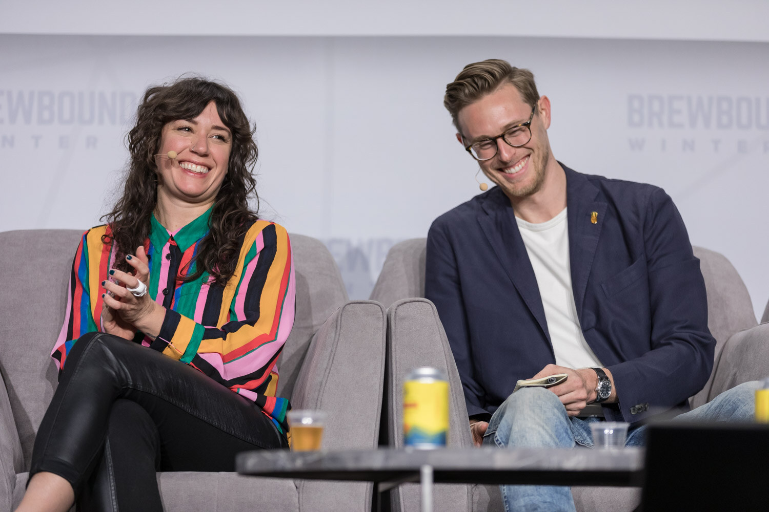 A woman in a colorful striped blouse and a man in a navy blazer laugh on stage with a can and a small beer glass on the table.