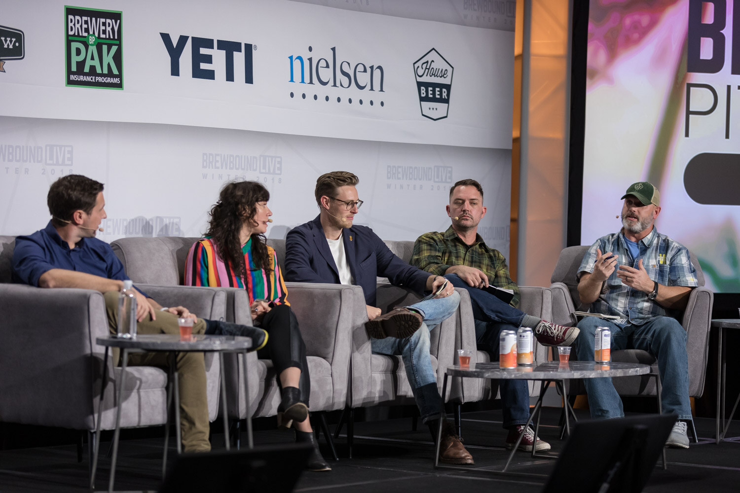 Five people on gray stage couches in front of Brewery Pak, YETI and Nielsen logos, with coffee tables and cans in front of them.