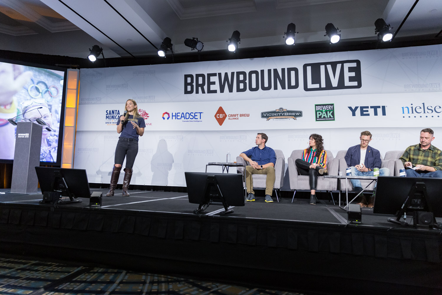 A wide view of the Brewbound Live stage with a woman presenting at left and four judges seated on couches at right.