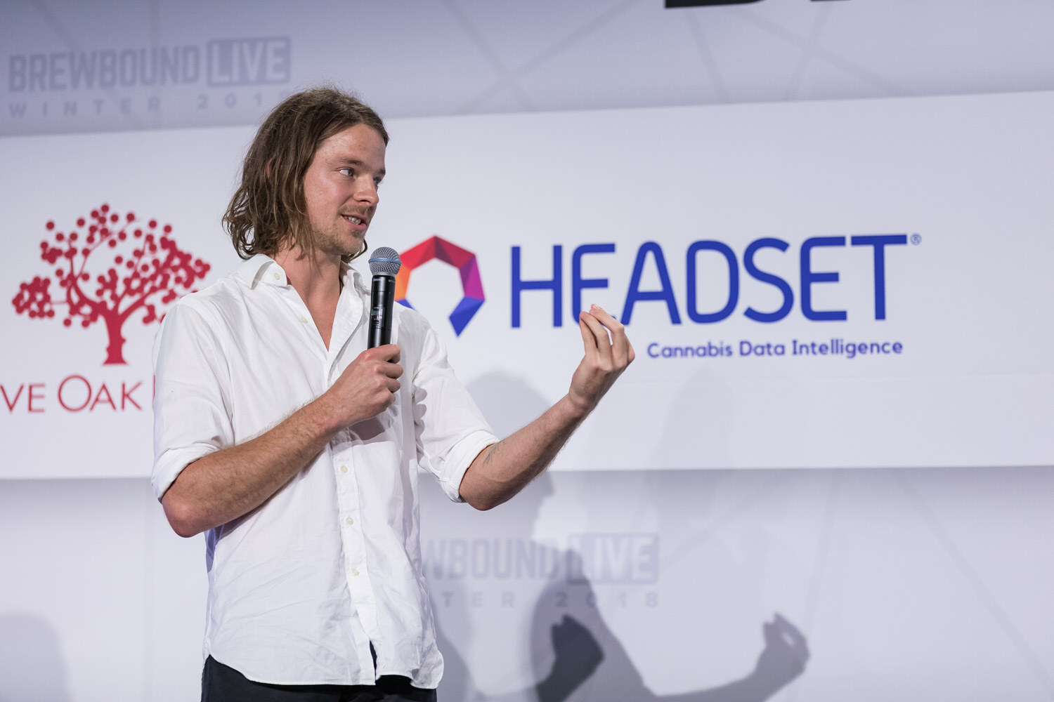 A man with long hair in a white shirt holds a microphone and gestures in front of a Headset logo.