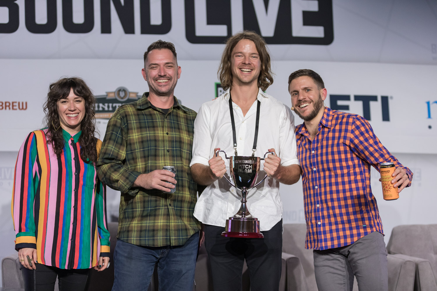 A woman in a striped blouse and three men on stage, the man in white holding up a silver trophy cup and the others holding cans.
