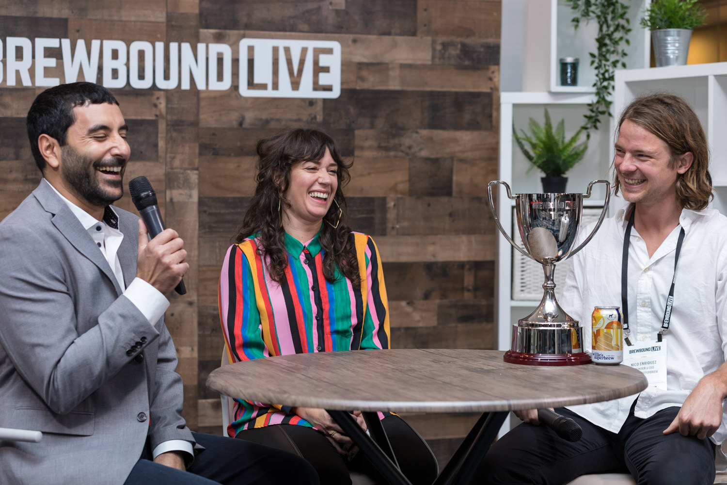 Three people laugh at a round table in front of a wood-paneled Brewbound Live wall, a silver trophy cup between them.