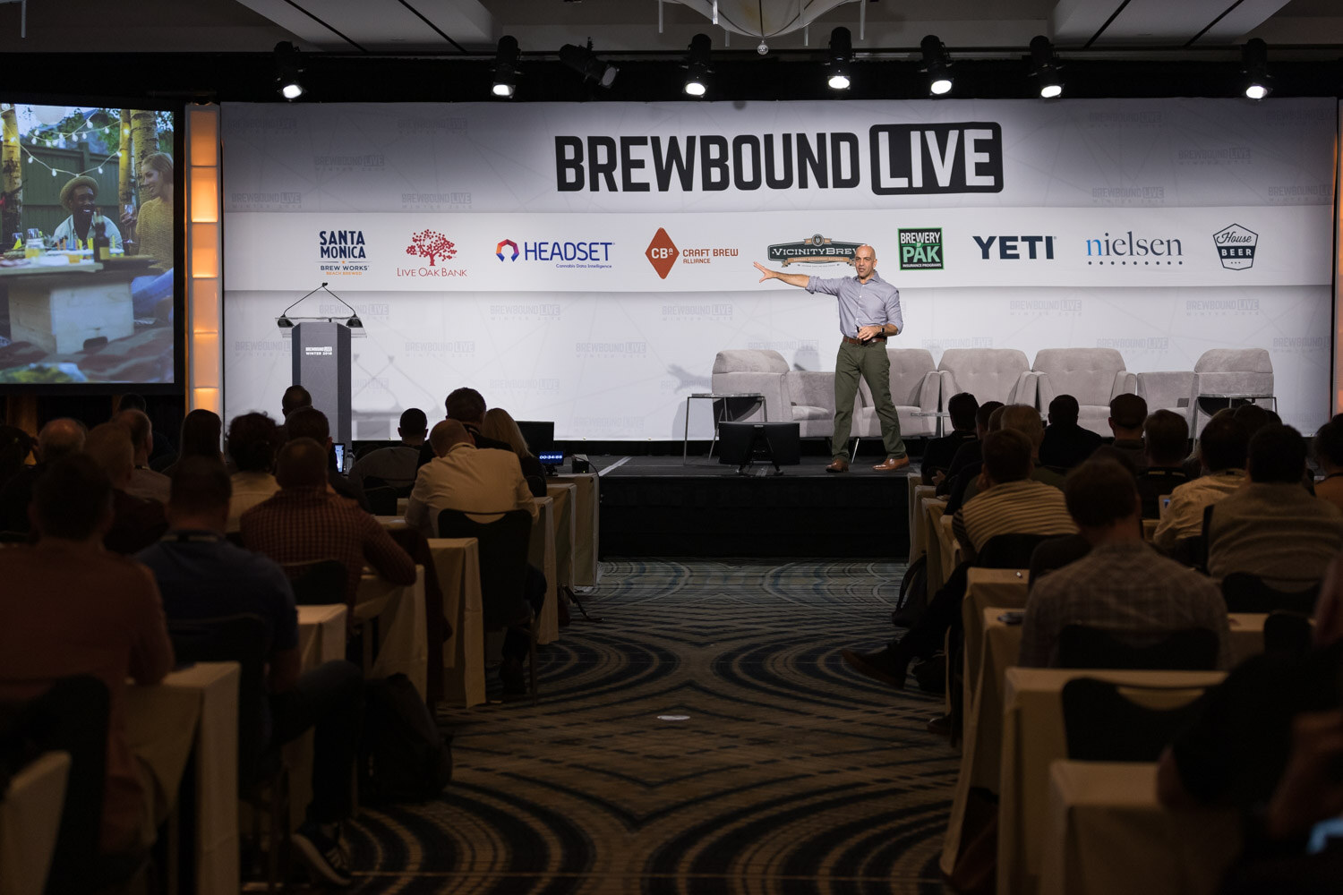 A man in a blue shirt speaks on a stage below a large Brewbound Live sponsor wall, with an audience at tables in the foreground.