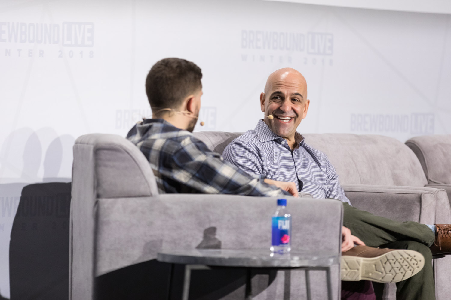 Two men sit on gray couches on stage, one in a plaid shirt holding papers and the other smiling widely.