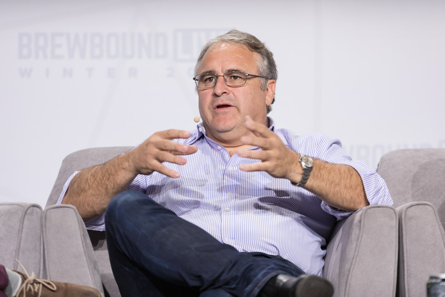 A man with glasses in a lavender button-up shirt leans back in an armchair and gestures while speaking.
