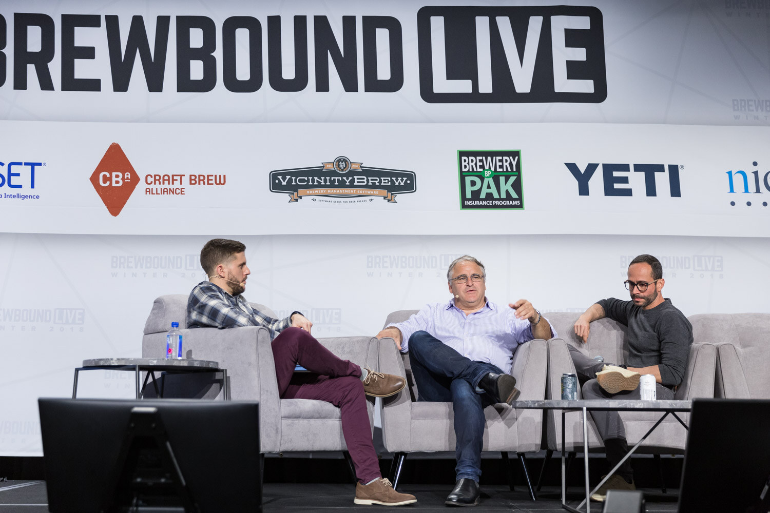 Three men on gray couches on stage in front of a Brewbound Live wall of sponsor logos.