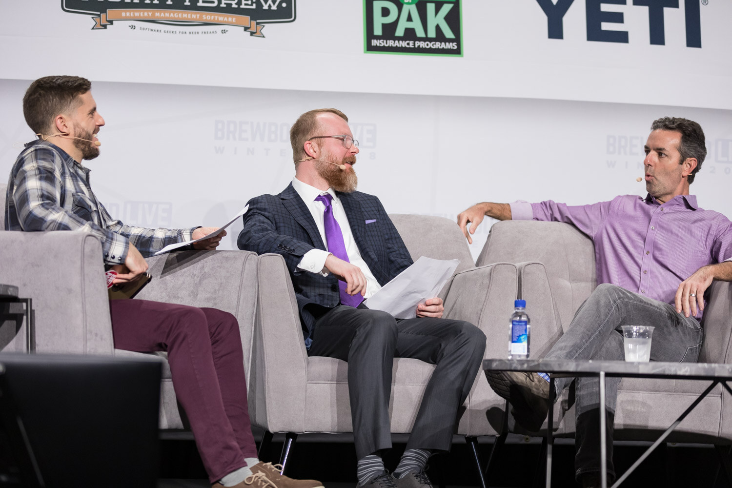 Three men on gray stage couches in front of a Brewbound Live sponsor wall, one holding papers.
