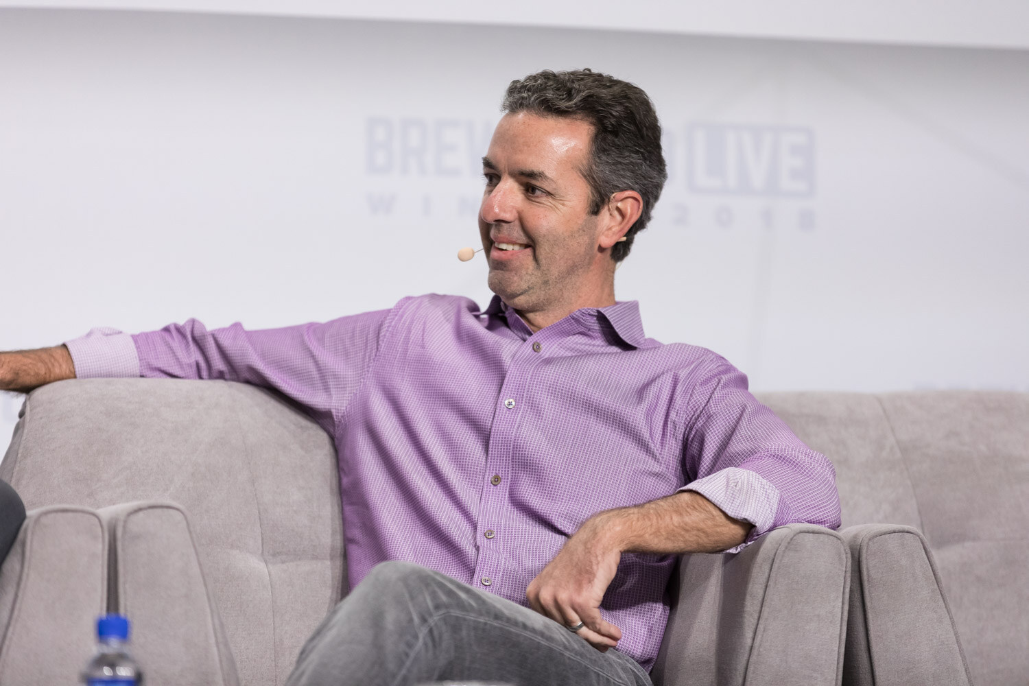 A man in a purple shirt relaxes in an armchair on stage with a water bottle in the foreground.