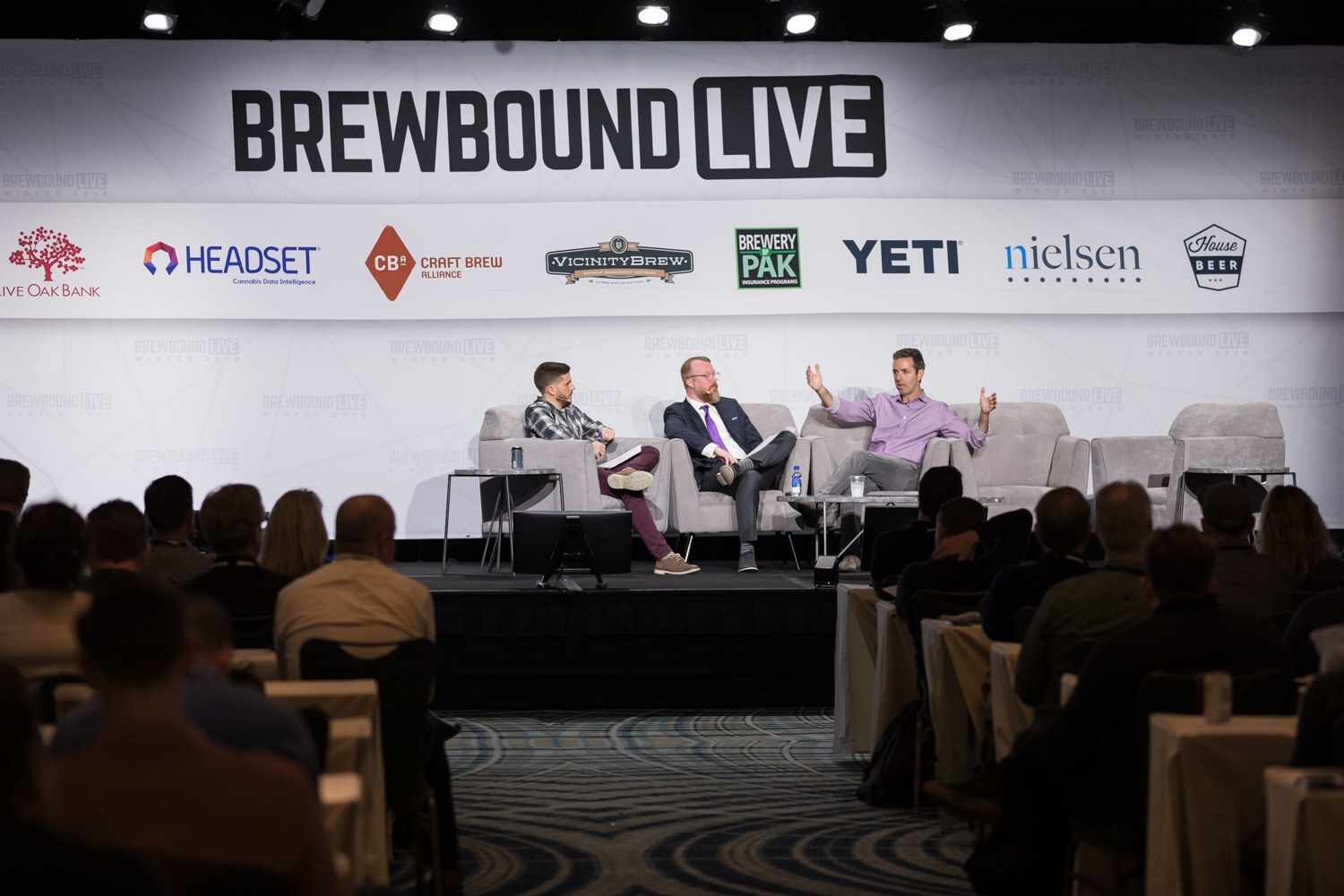 Three people on stage couches under a large Brewbound Live sign with sponsor logos, audience tables in the foreground.
