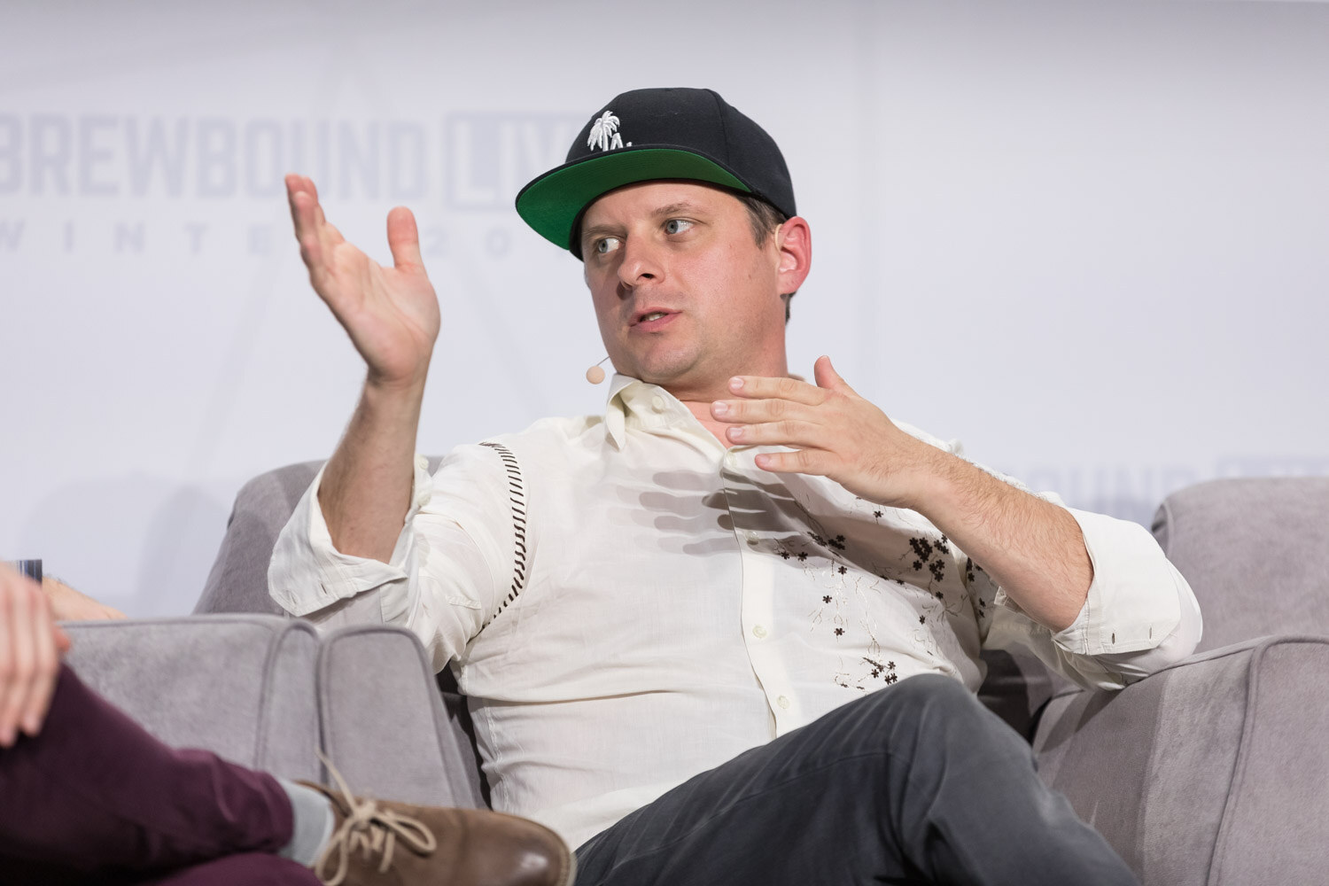 A man in a black and green ball cap and white patterned shirt gestures as he talks in an armchair.