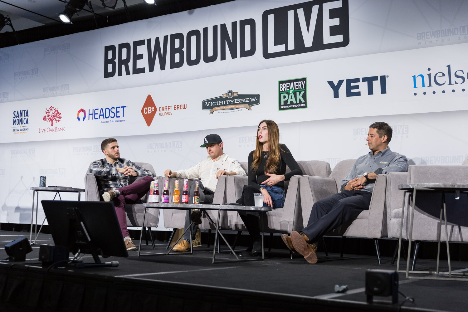 Four people on gray couches on stage with several brightly labeled bottles on a low table.