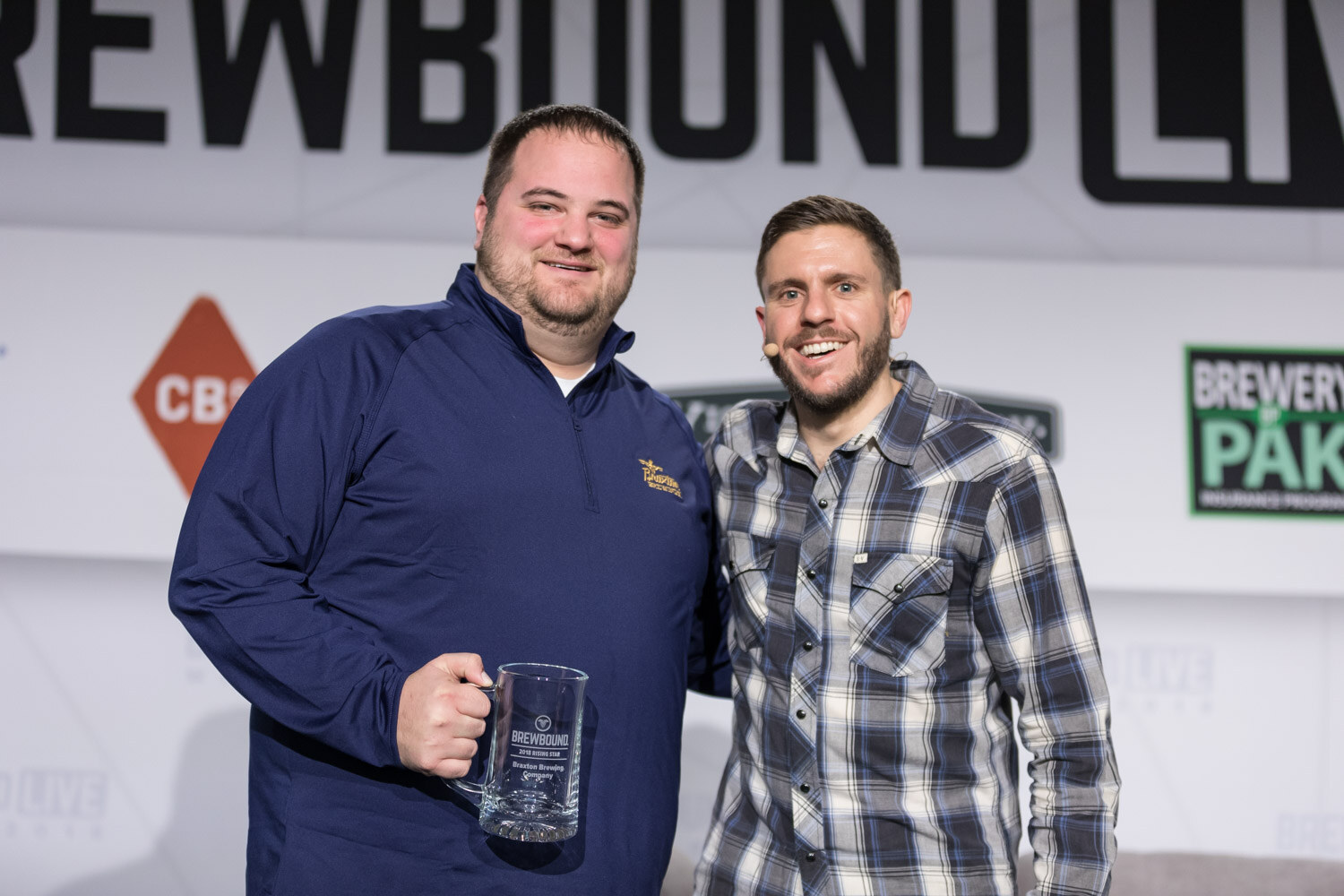 Two smiling men pose for a photo, one in a navy pullover holding an engraved glass mug and the other in a plaid shirt with a headset microphone.