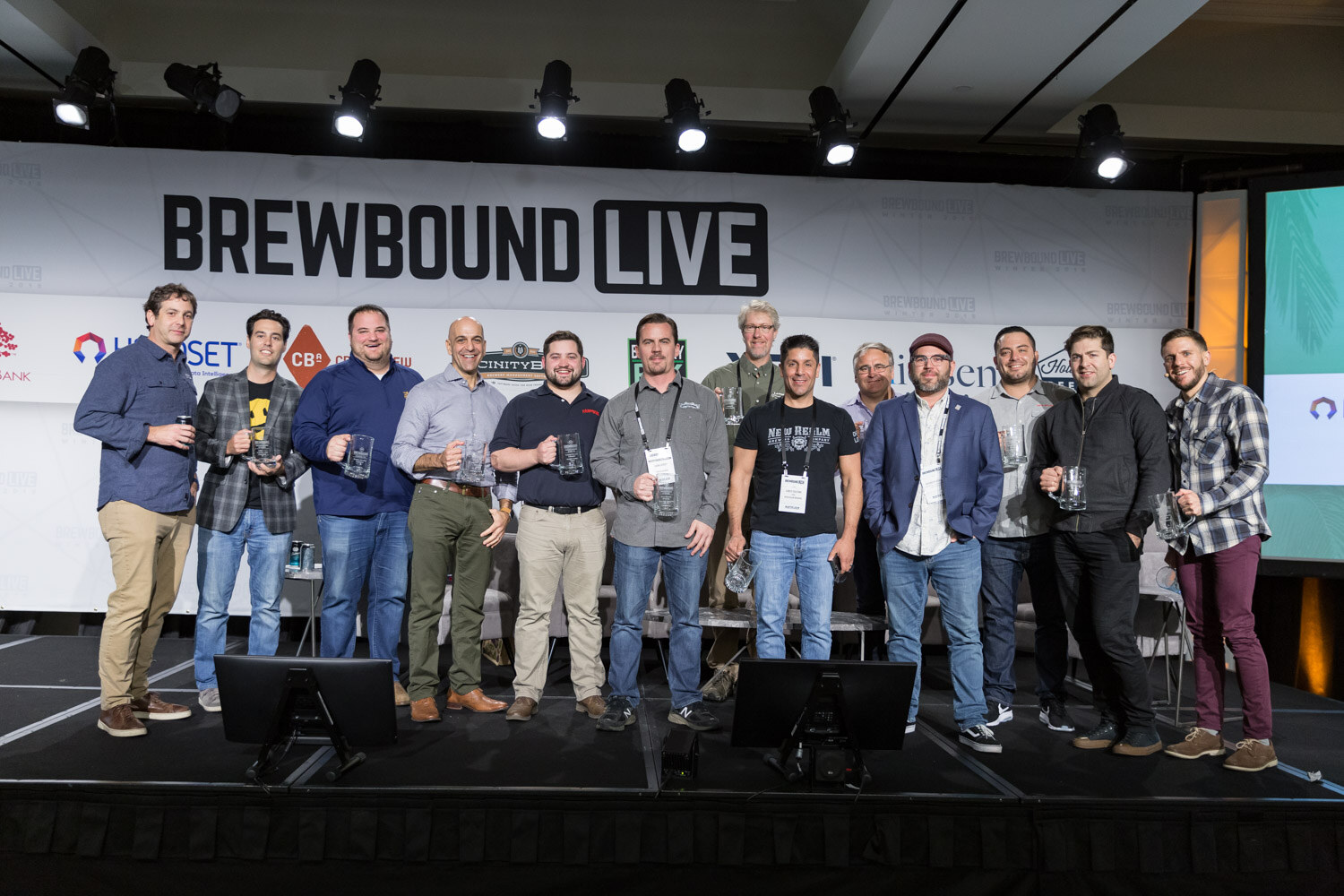 A line of about a dozen people holding glass mugs on a stage in front of a Brewbound Live sign.