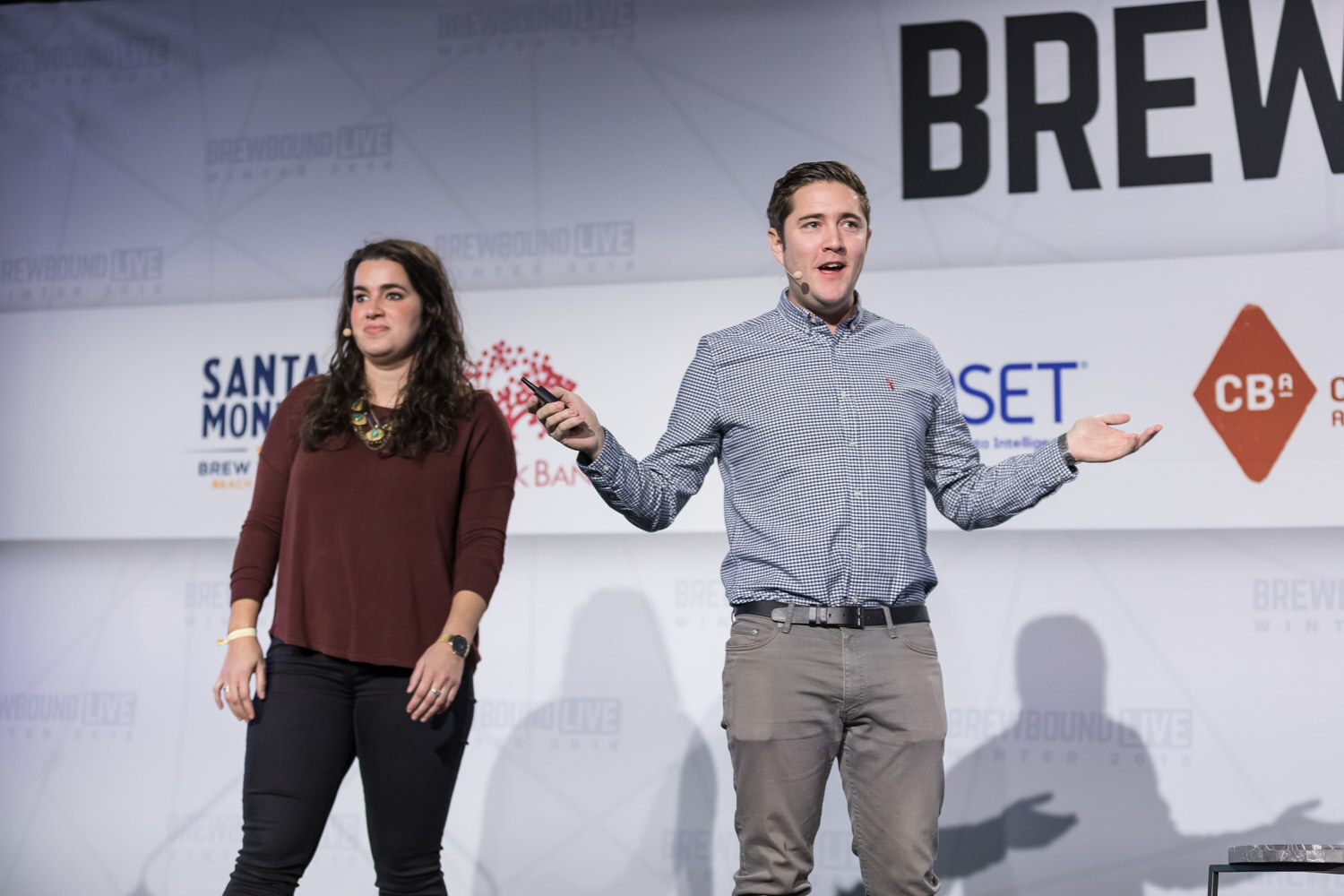 A woman in a maroon top and a man in a blue checked shirt stand on stage in front of a Headset sponsor logo.