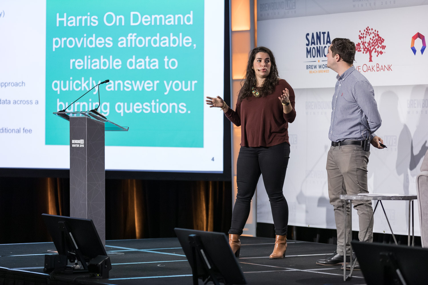 A woman and a man on stage beside a podium and a large teal slide that begins Harris On Demand provides affordable, reliable data.
