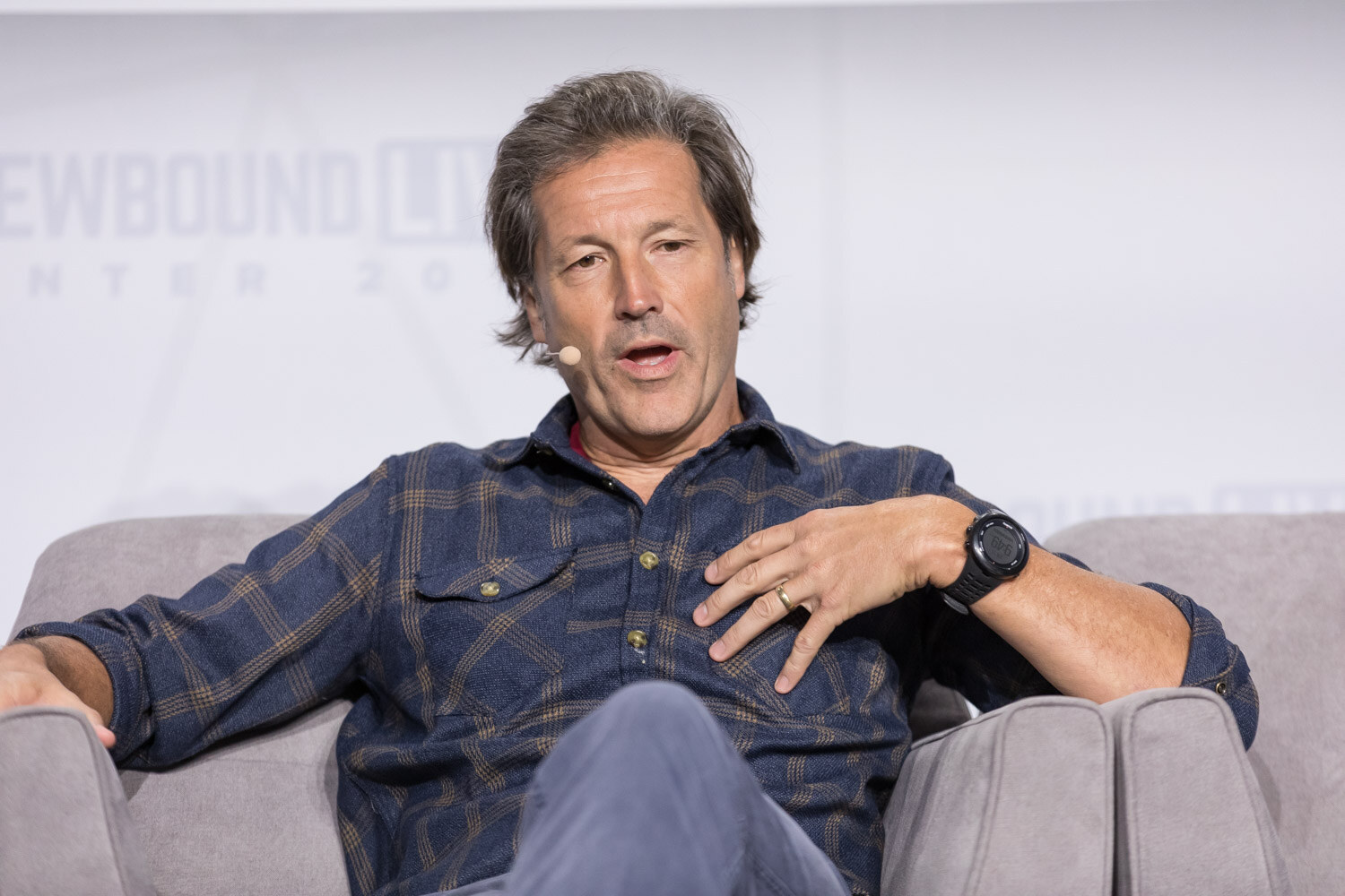 A man in a dark plaid shirt with a headset microphone gestures while seated in an armchair.