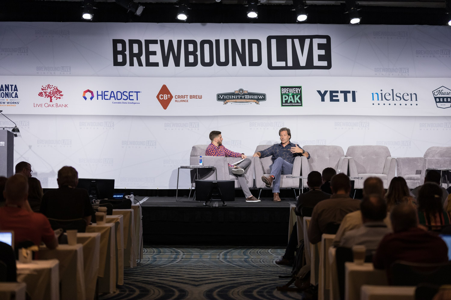Two people on stage couches under a large Brewbound Live banner, audience tables in the foreground.