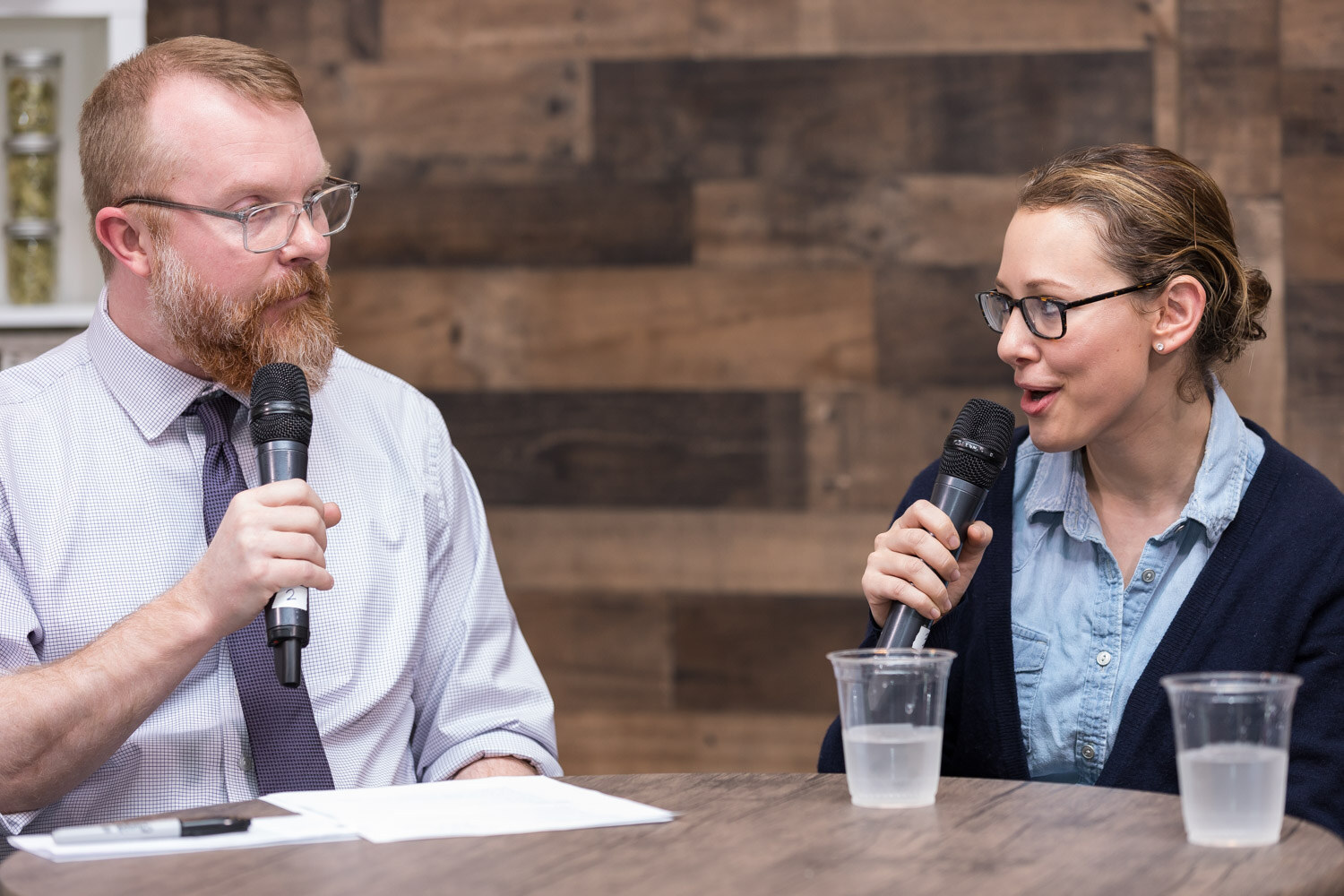 A man in a tie holding a microphone and a woman in a denim shirt holding a microphone at a round table.