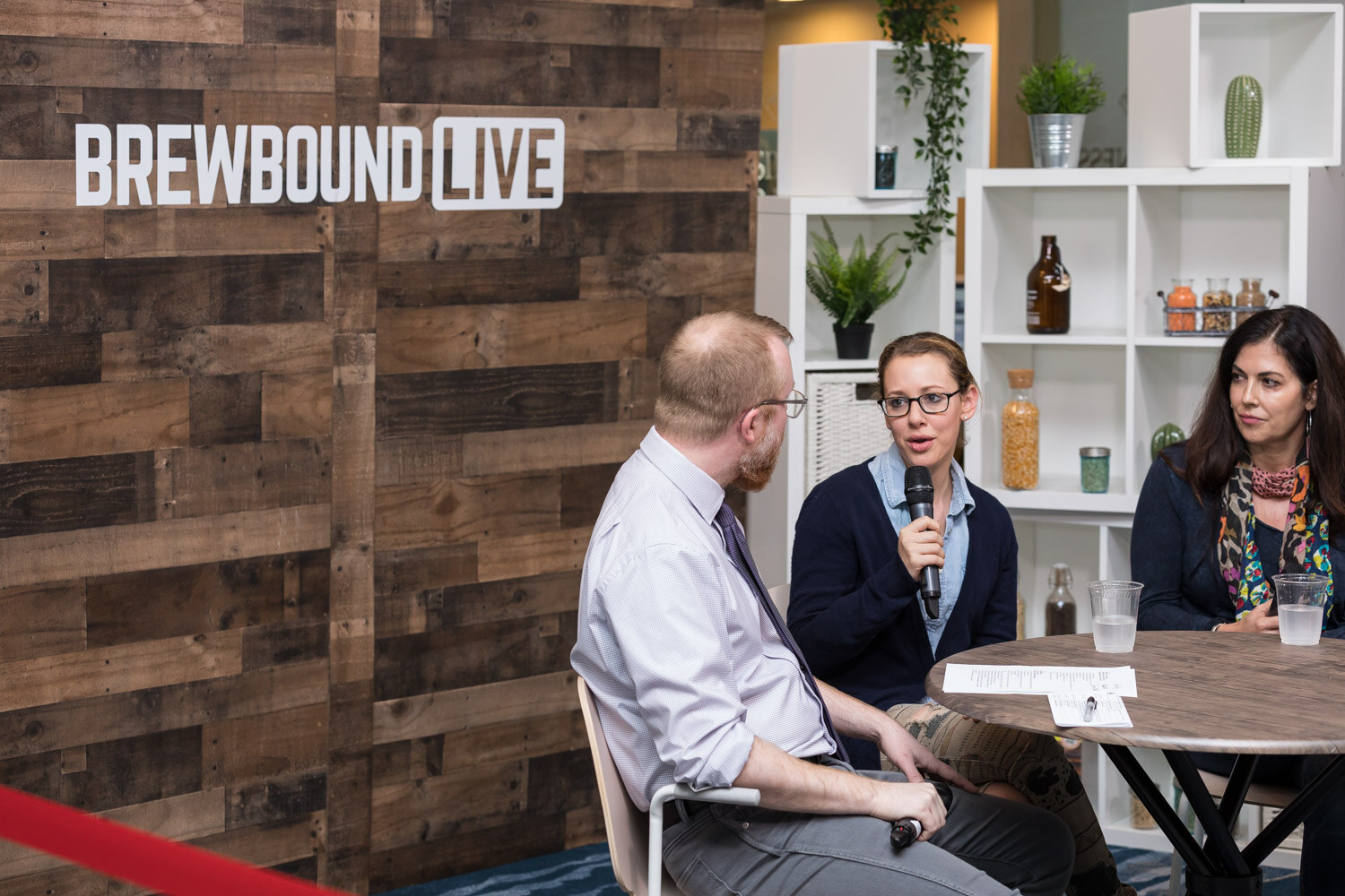 Three people seated around a small table in front of a wood-paneled wall with a Brewbound Live logo and white shelves.