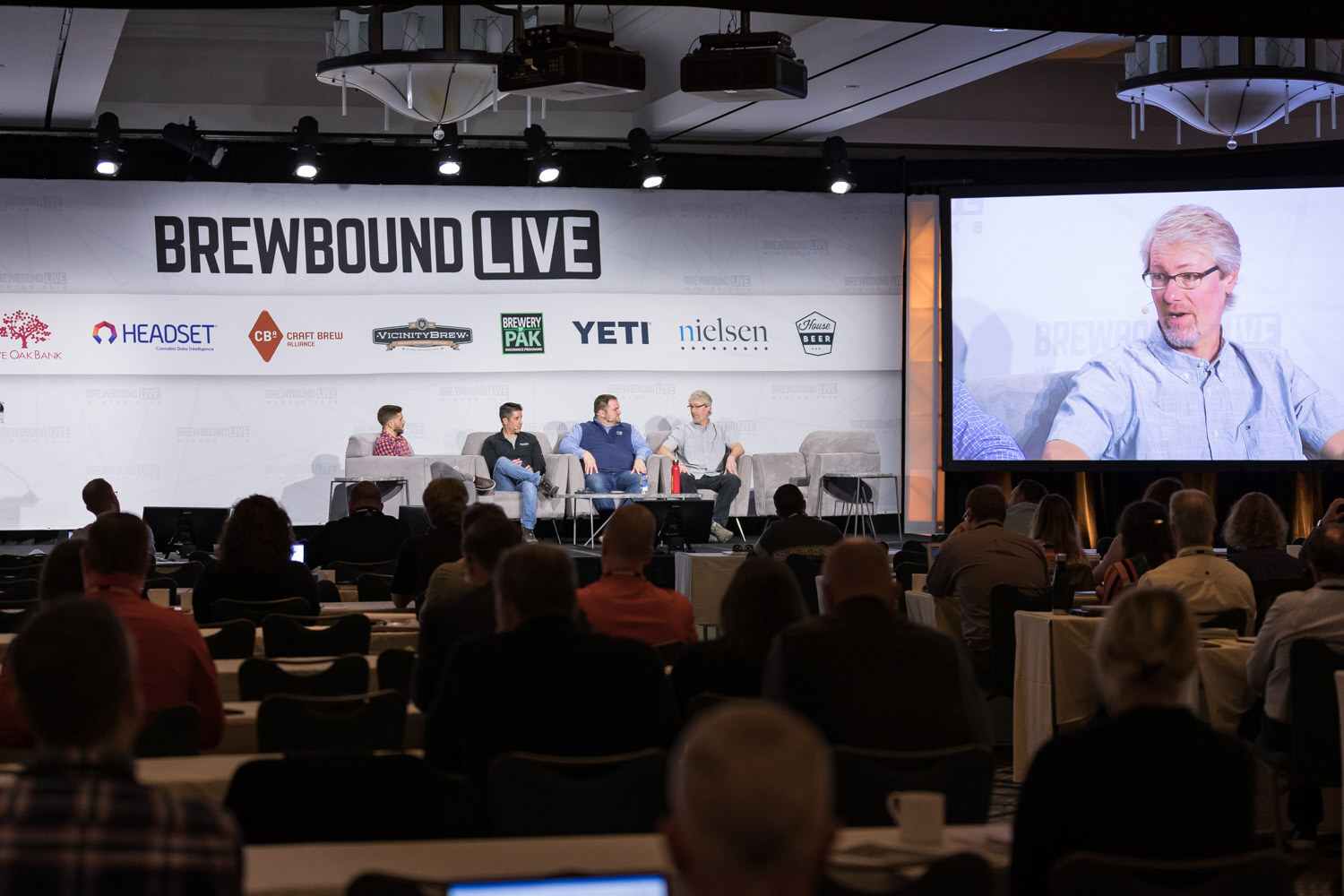 A wide view of four people on stage couches, a big screen at right showing a close-up of one panelist, and audience tables in front.