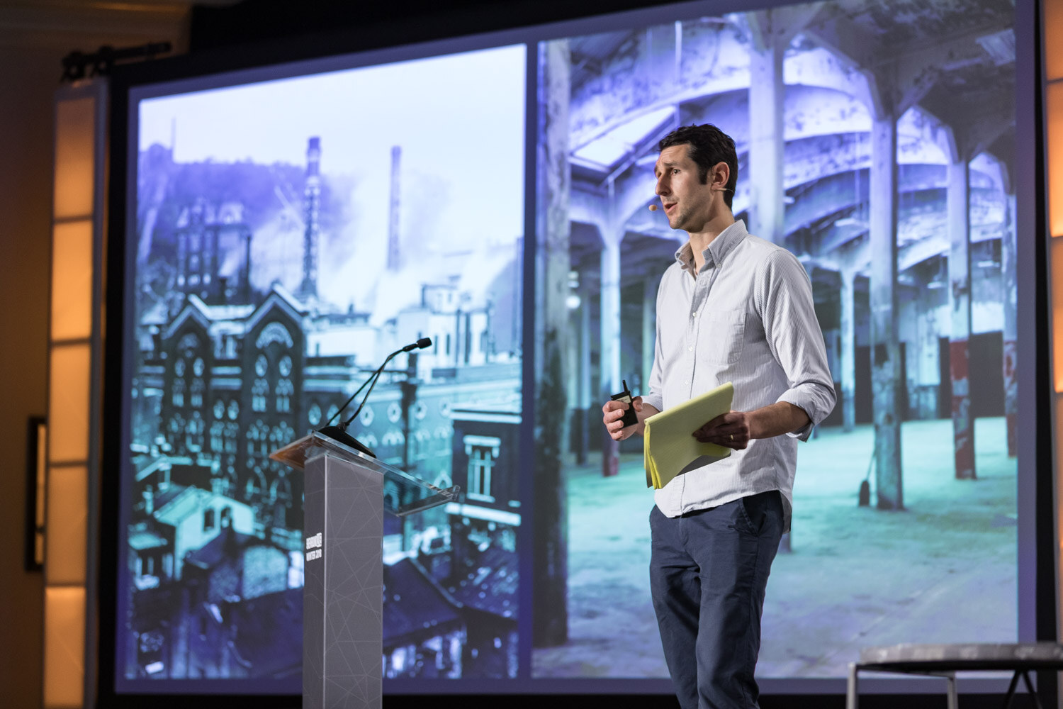 A man beside a podium in front of a large screen showing two images of historic industrial brewery buildings.