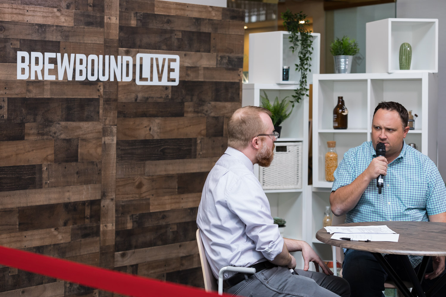 Two men with microphones at a round table in front of a wooden wall with the Brewbound Live logo.