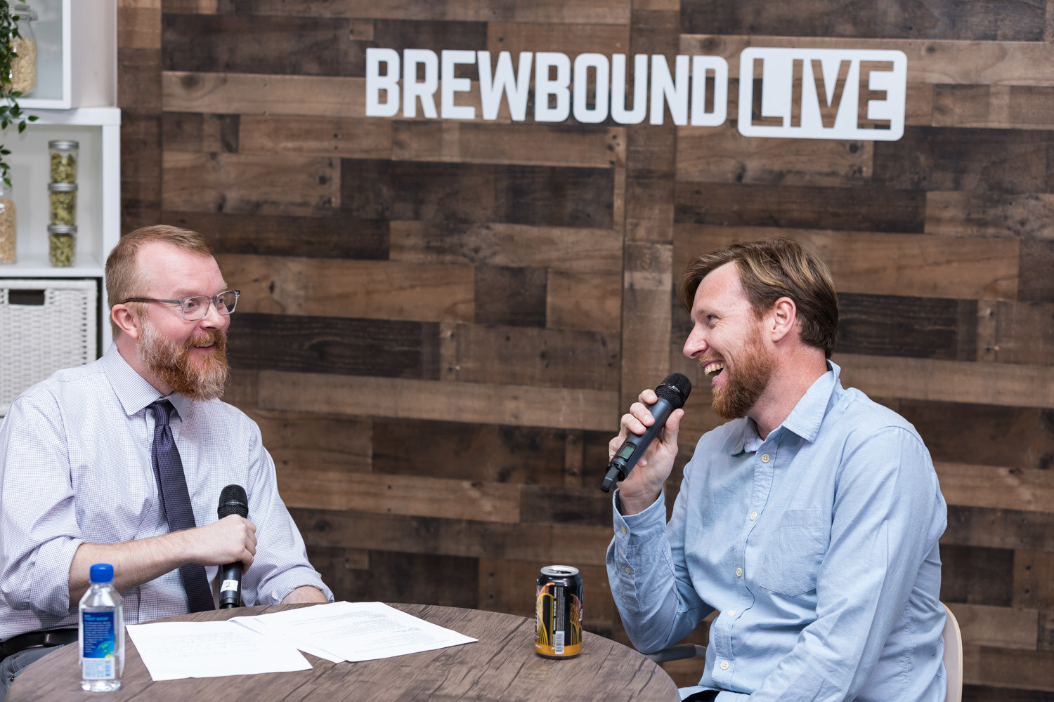 A man in a tie and a bearded man in a light blue shirt laugh across a table with microphones, a can and a water bottle.