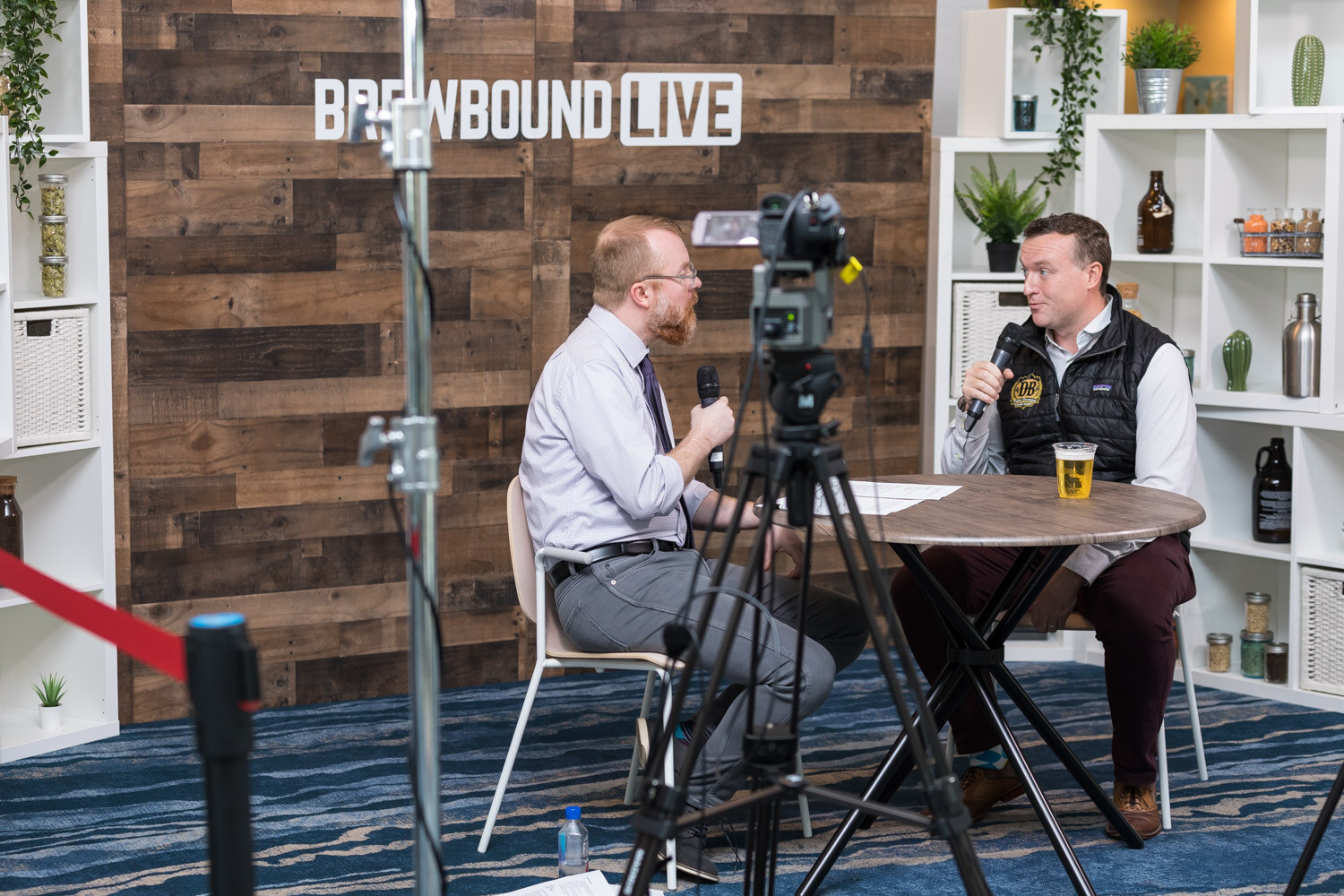 A camera in the foreground records two men in conversation at a small table with a pint of beer, in front of a Brewbound Live sign.