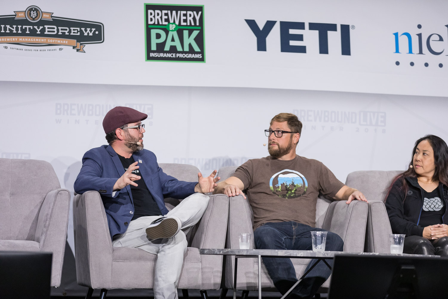 A man in a maroon flat cap and blue jacket, a bearded man in a brown T-shirt and a woman in black sit on stage couches.