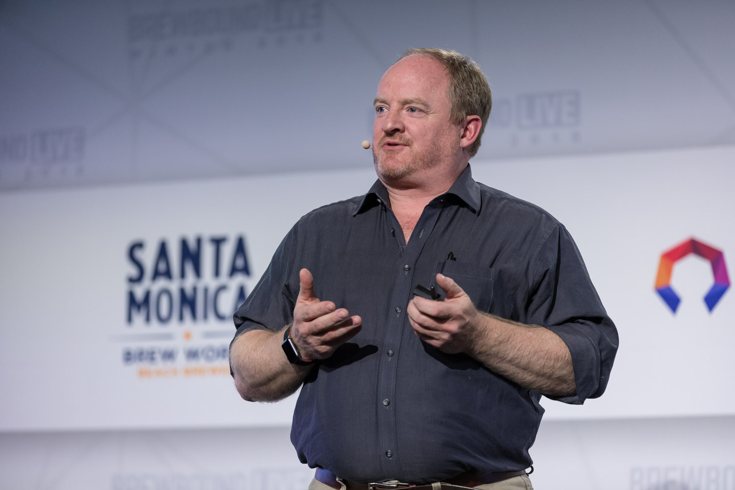 A man in a dark gray shirt with a headset microphone gestures with both hands in front of a Santa Monica Brew Works logo.