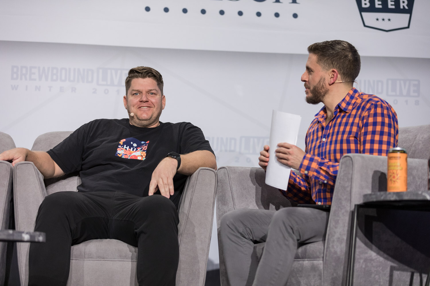A man in a black T-shirt and another in an orange plaid shirt sit across from each other on gray armchairs on stage.