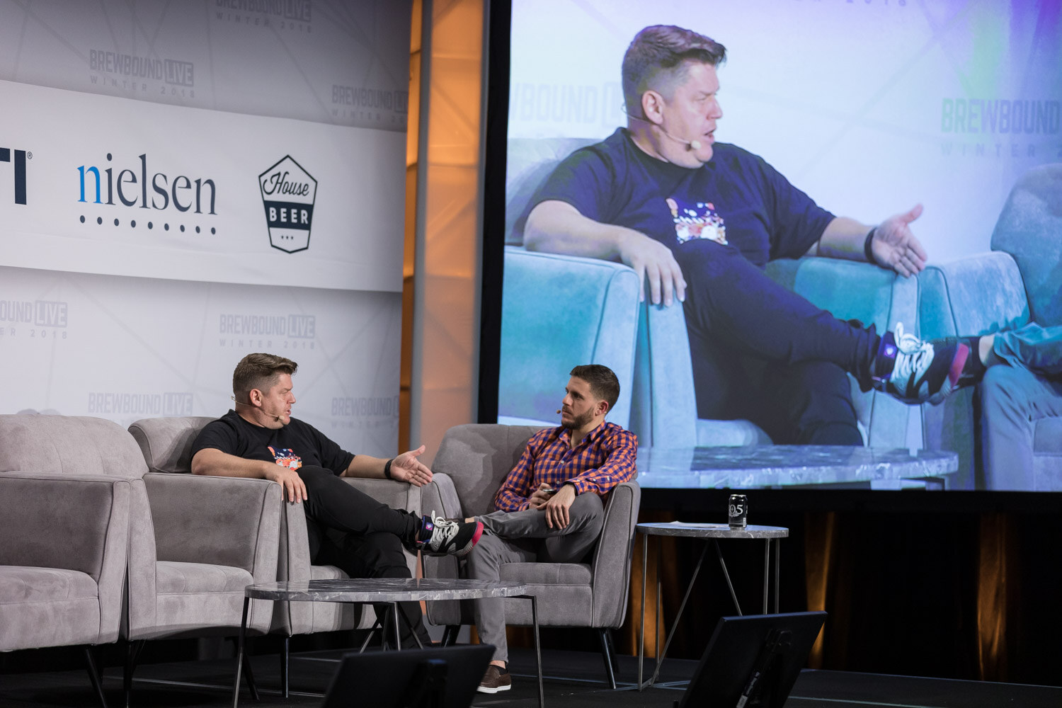 Two people seated on stage with a large screen behind showing a close-up of the same conversation.