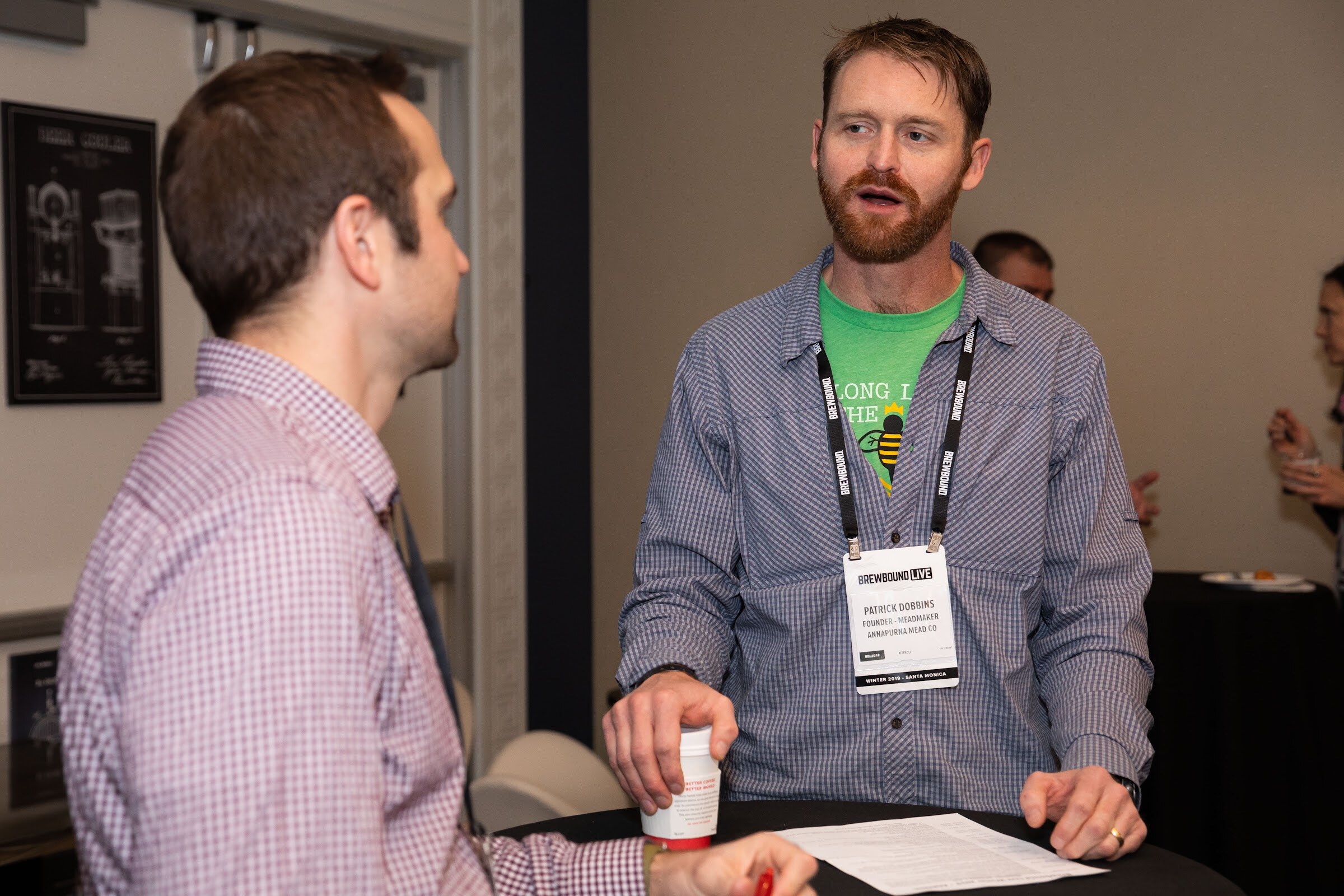 Two men with badges talking at a high-top table with coffee cups; one wears a green bee T-shirt.