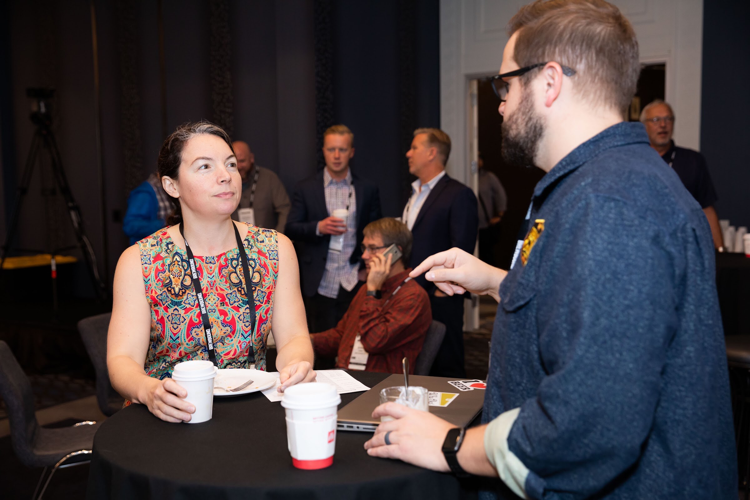 Woman in a colorful sleeveless dress holding a coffee cup while a bearded man in a denim shirt points.