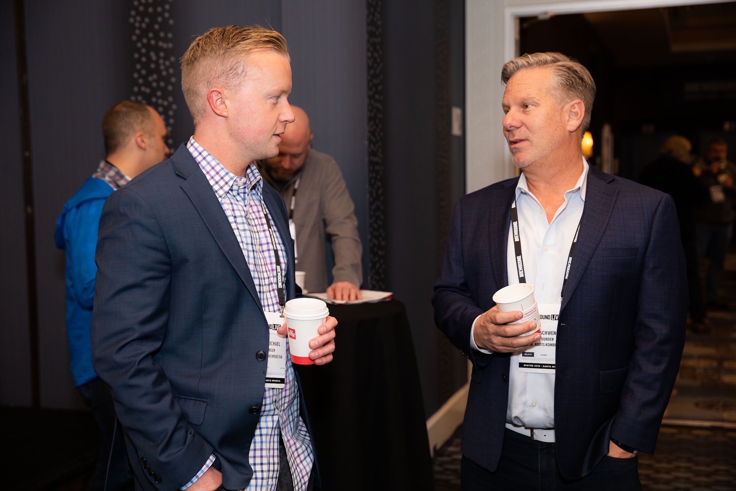 Two men in blazers holding paper coffee cups in a hallway.